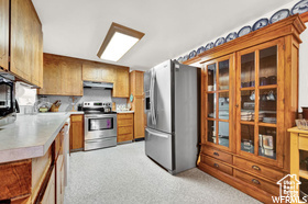 Kitchen with stainless steel appliances, under cabinet range hood, light countertops, brown cabinetry, and glass insert cabinets