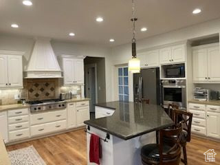 Kitchen with custom exhaust hood, stainless steel appliances, white cabinets, a breakfast bar, and light wood-style flooring