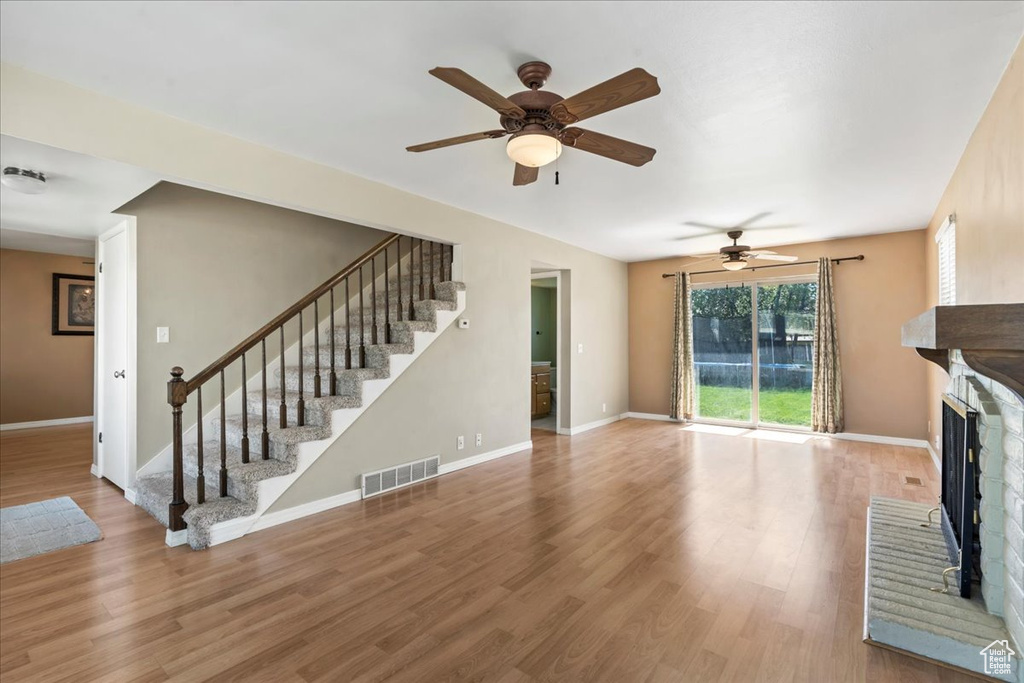 Unfurnished living room featuring a ceiling fan, stairs, a brick fireplace, and wood finished floors