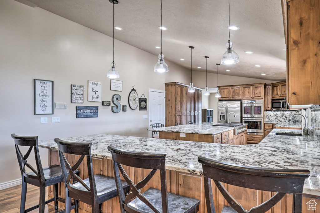 Kitchen with a kitchen island with sink, stainless steel appliances, brown cabinetry, wood finished floors, and light stone counters