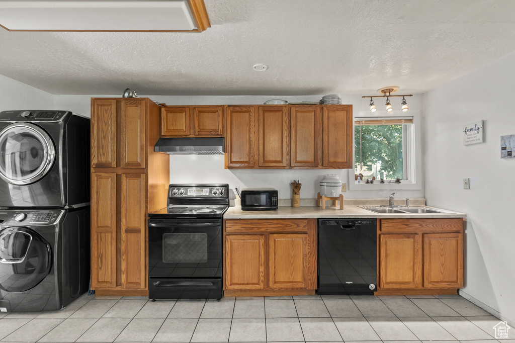 Kitchen with black appliances, estacked washer and dryer, brown cabinetry, light countertops, and under cabinet range hood