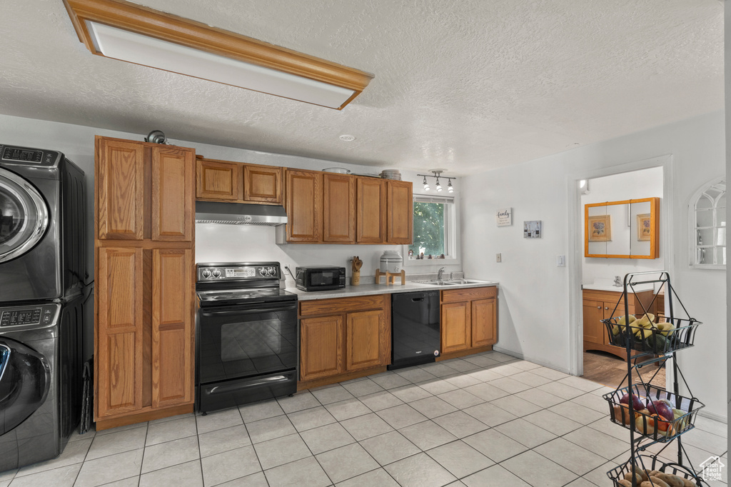 Kitchen featuring black appliances, under cabinet range hood, stacked washer / drying machine, brown cabinets, and a textured ceiling