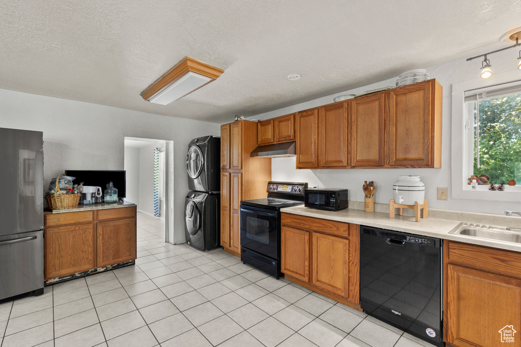 Kitchen with black appliances, stacked washer and clothes dryer, under cabinet range hood, brown cabinetry, and a textured ceiling