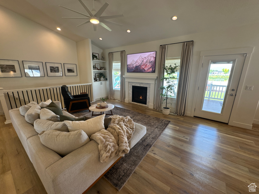 Living area with a warm lit fireplace, ceiling fan, healthy amount of natural light, lofted ceiling, and wood finished floors