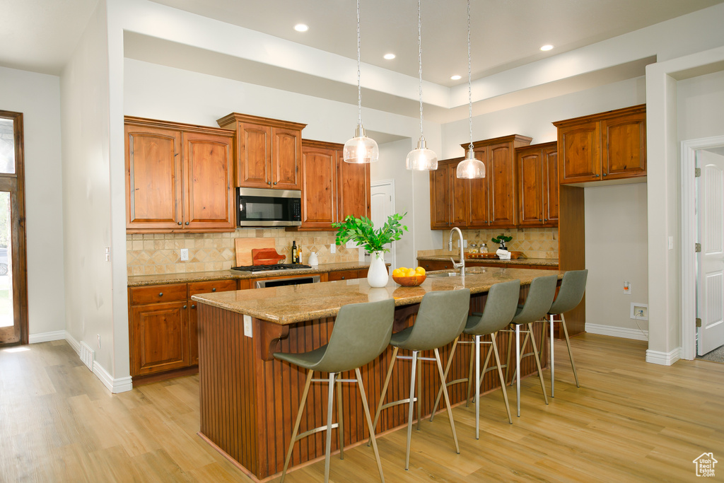 Kitchen featuring stainless steel microwave, light wood-type flooring, brown cabinetry, a kitchen island with sink, and recessed lighting