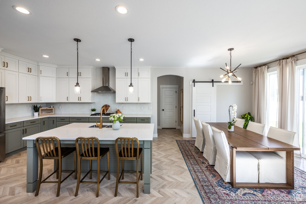 Kitchen featuring wall chimney range hood, a barn door, gray cabinets, light countertops, and a center island with sink