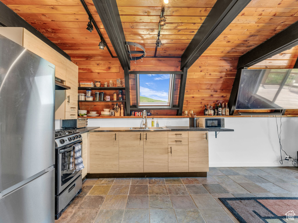 Kitchen with track lighting, appliances with stainless steel finishes, wooden walls, light brown cabinets, and stone tile floors