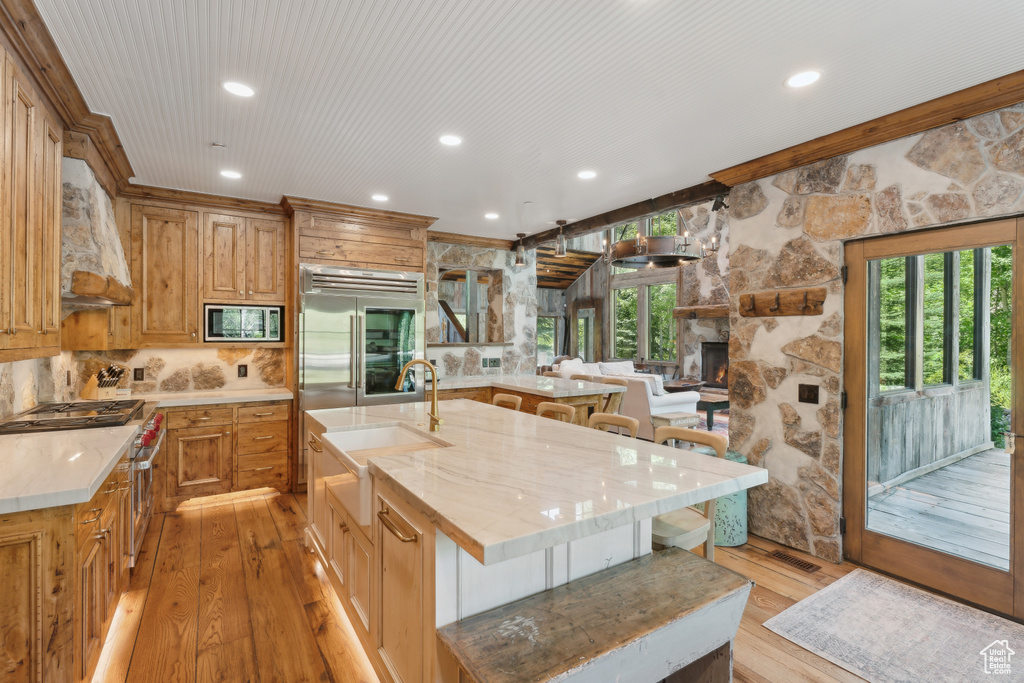 Kitchen with built in appliances, premium range hood, light wood-type flooring, a kitchen breakfast bar, and a stone fireplace