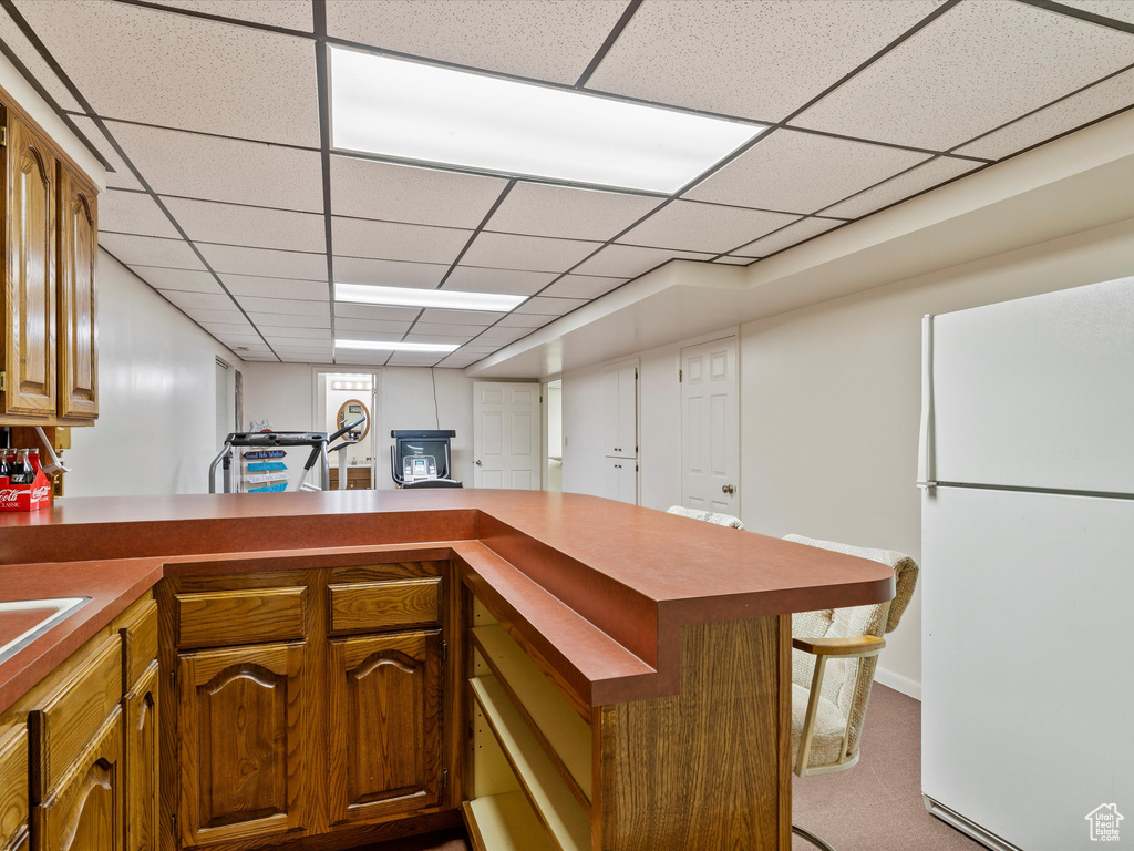 Kitchen featuring freestanding refrigerator, a peninsula, a paneled ceiling, light carpet, and brown cabinetry
