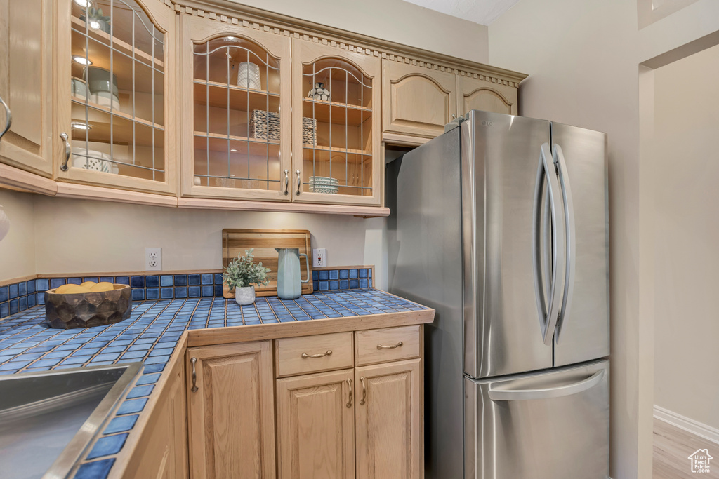 Kitchen featuring freestanding refrigerator, tile counters, glass insert cabinets, and light brown cabinetry