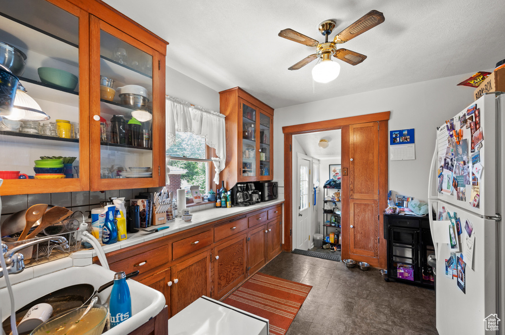 Kitchen with freestanding refrigerator, a ceiling fan, glass insert cabinets, brown cabinets, and tile countertops