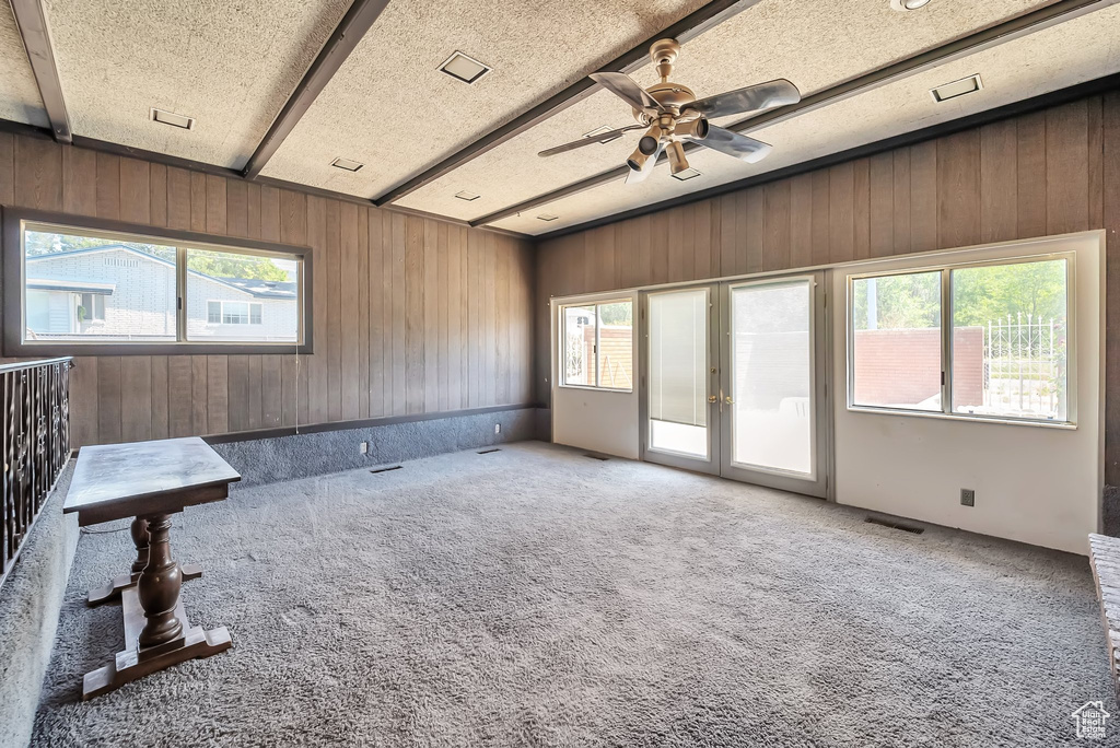 Carpeted spare room featuring wooden walls, healthy amount of natural light, a ceiling fan, a textured ceiling, and beam ceiling