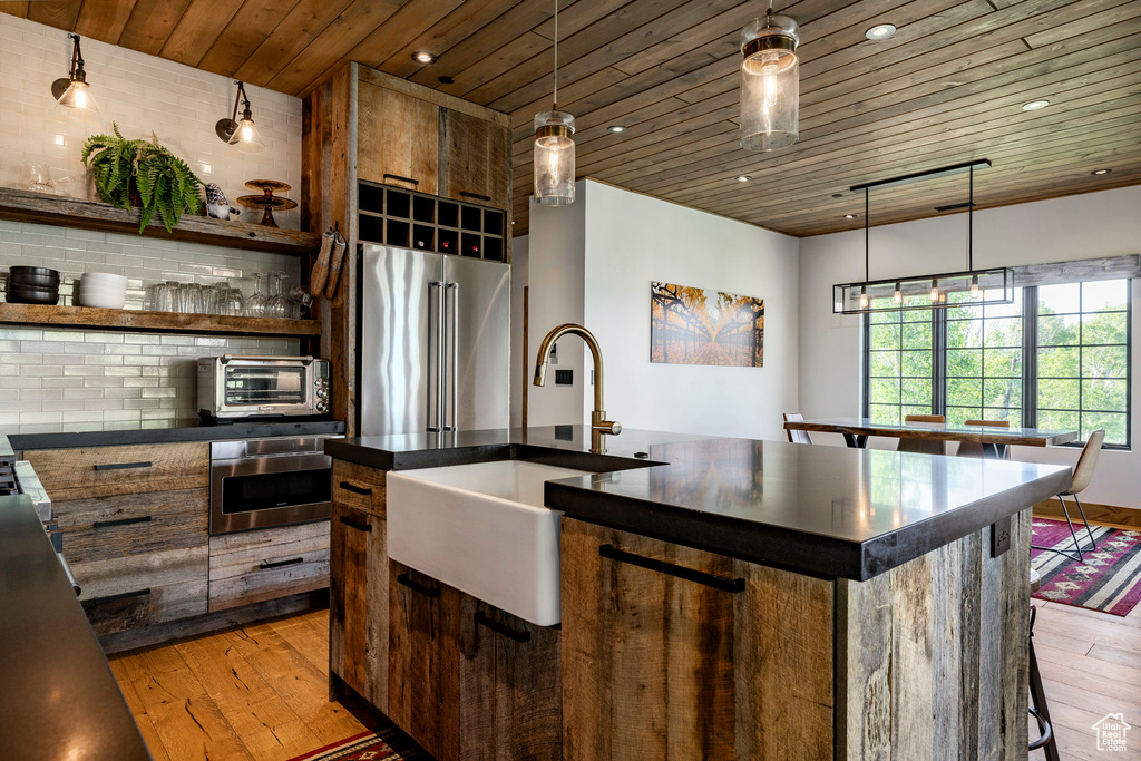 Kitchen featuring appliances with stainless steel finishes, light wood-type flooring, dark countertops, wood ceiling, and a kitchen island with sink
