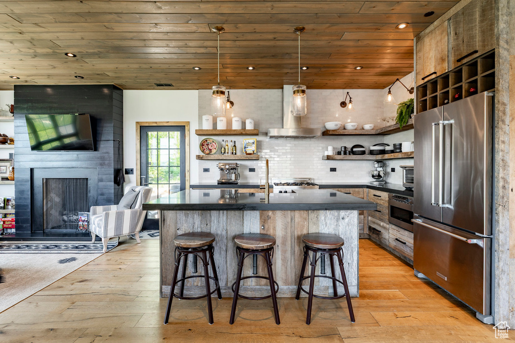 Kitchen with high end refrigerator, open shelves, wood ceiling, dark countertops, and wall chimney range hood