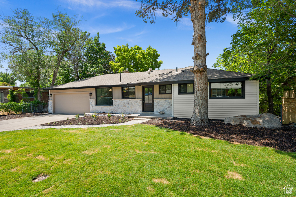 View of front of house featuring stone siding, a garage, driveway, a front lawn, and a shingled roof
