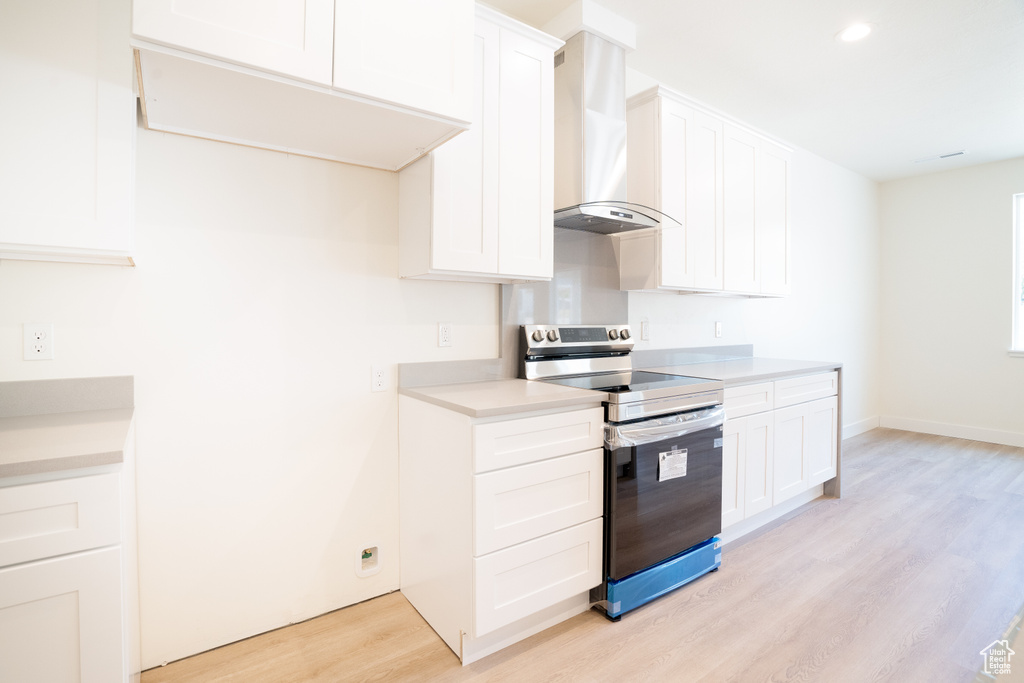 Kitchen with stainless steel range with electric stovetop, wall chimney exhaust hood, light wood-style flooring, light countertops, and white cabinetry