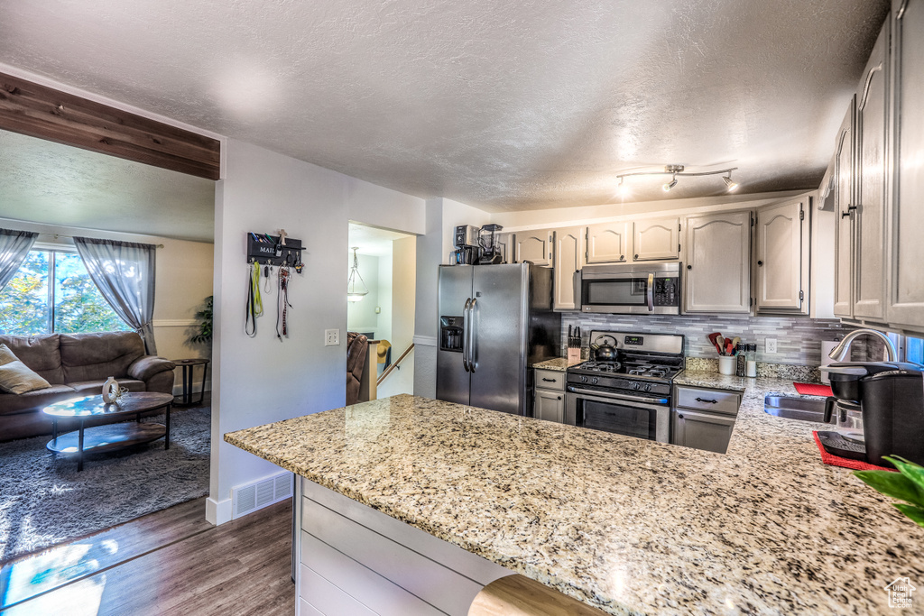 Kitchen with appliances with stainless steel finishes, a peninsula, a textured ceiling, dark wood-type flooring, and light stone counters