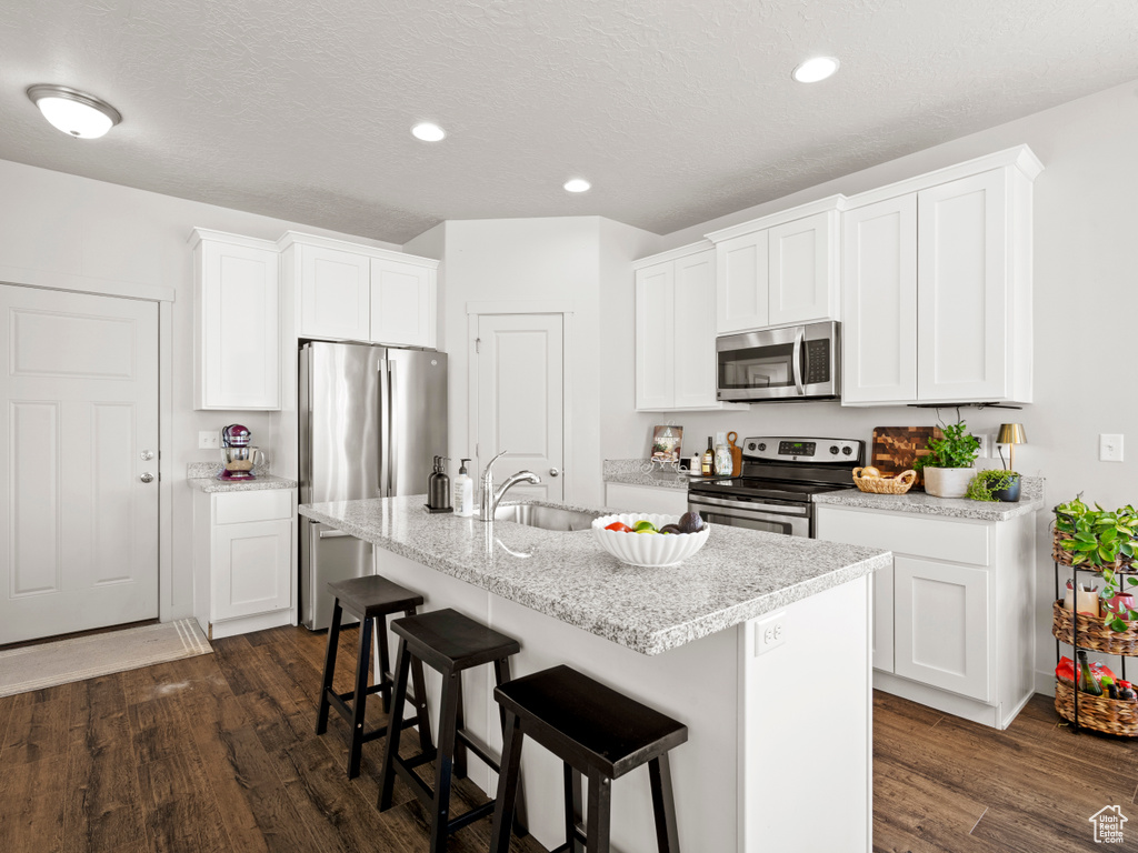 Kitchen with appliances with stainless steel finishes, dark wood finished floors, a kitchen breakfast bar, an island with sink, and white cabinets