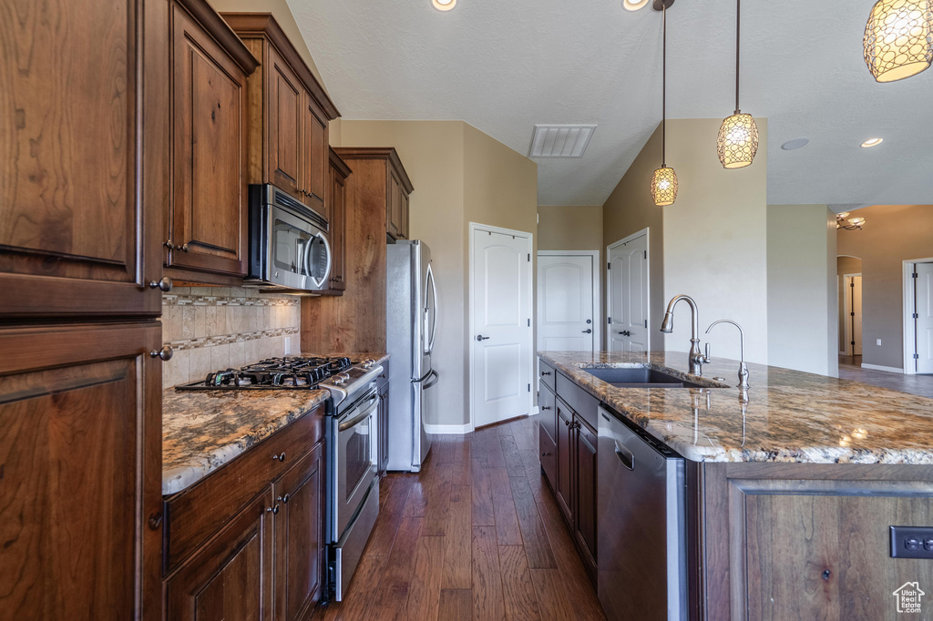 Kitchen with stainless steel appliances, dark wood-type flooring, dark stone counters, a center island with sink, and pendant lighting