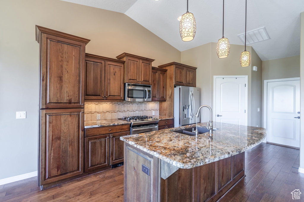 Kitchen with appliances with stainless steel finishes, dark wood-type flooring, an island with sink, decorative backsplash, and light stone countertops