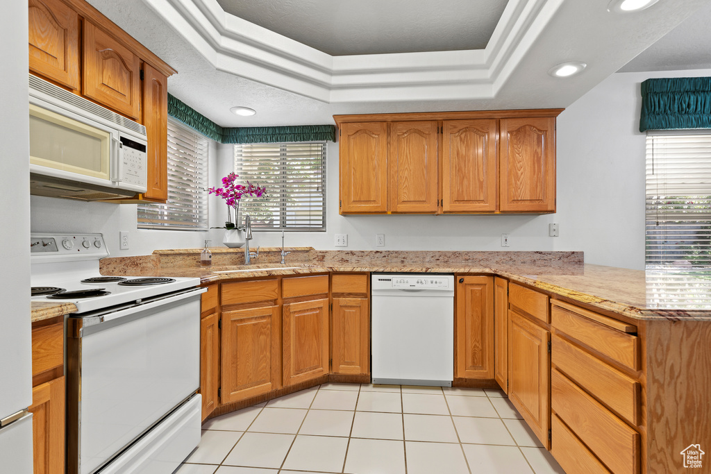 Kitchen featuring white appliances, a raised ceiling, a peninsula, a sink, and plenty of natural light