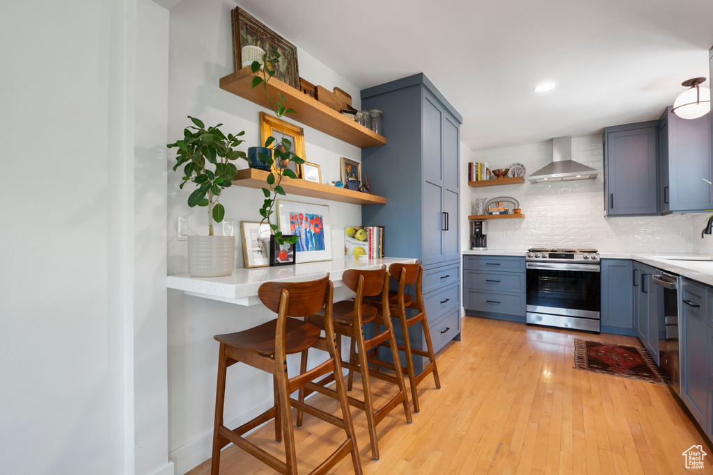 Kitchen with open shelves, stainless steel stove, wall chimney range hood, decorative backsplash, and light wood-style floors