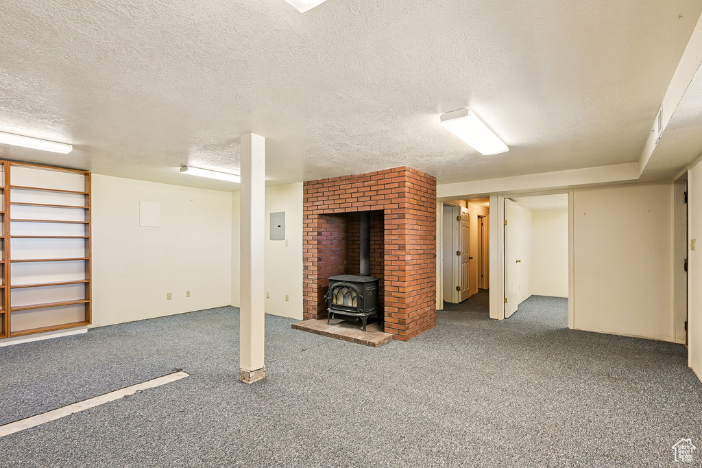 Finished basement with a wood stove, carpet flooring, a textured ceiling, and electric panel