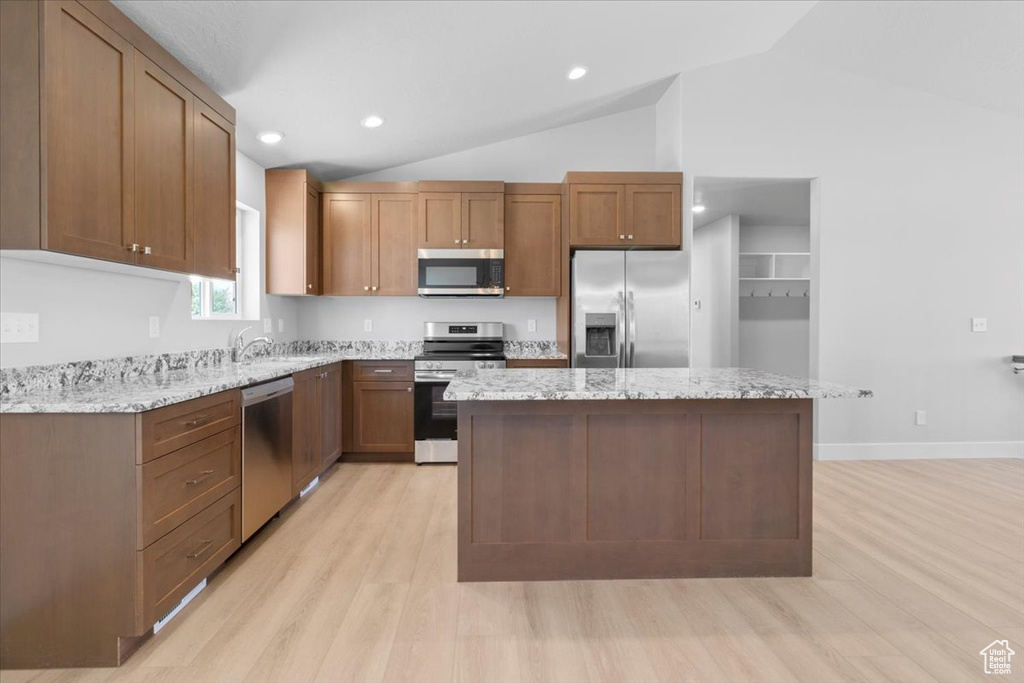 Kitchen with stainless steel appliances, light wood-style flooring, vaulted ceiling, a kitchen island, and light stone counters