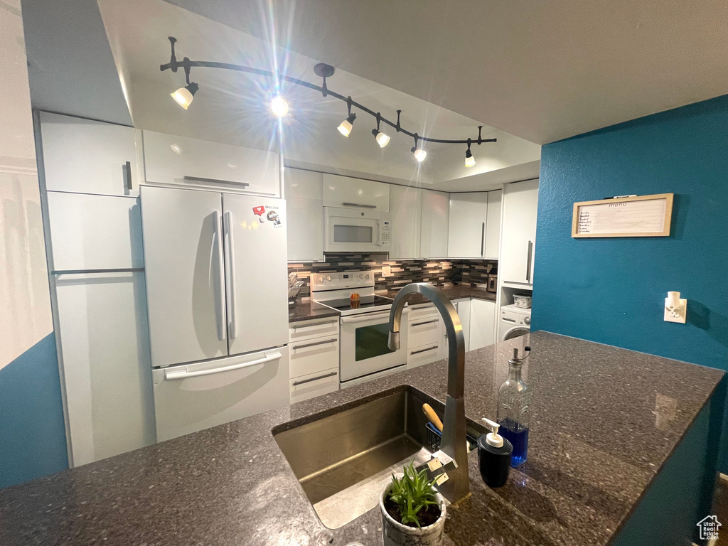 Kitchen with white appliances, rail lighting, white cabinets, tasteful backsplash, and dark stone countertops