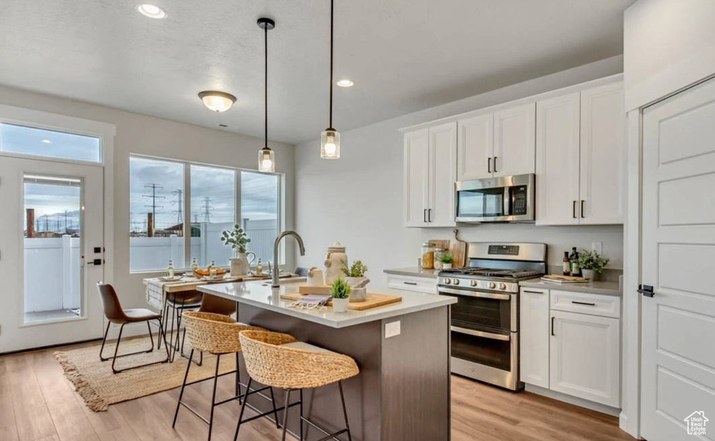 Kitchen featuring stainless steel appliances, an island with sink, light wood finished floors, light countertops, and white cabinets