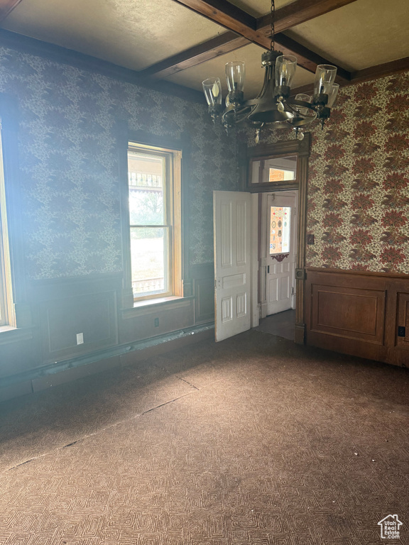 Empty room featuring wallpapered walls, a wainscoted wall, a chandelier, carpet flooring, and beamed ceiling
