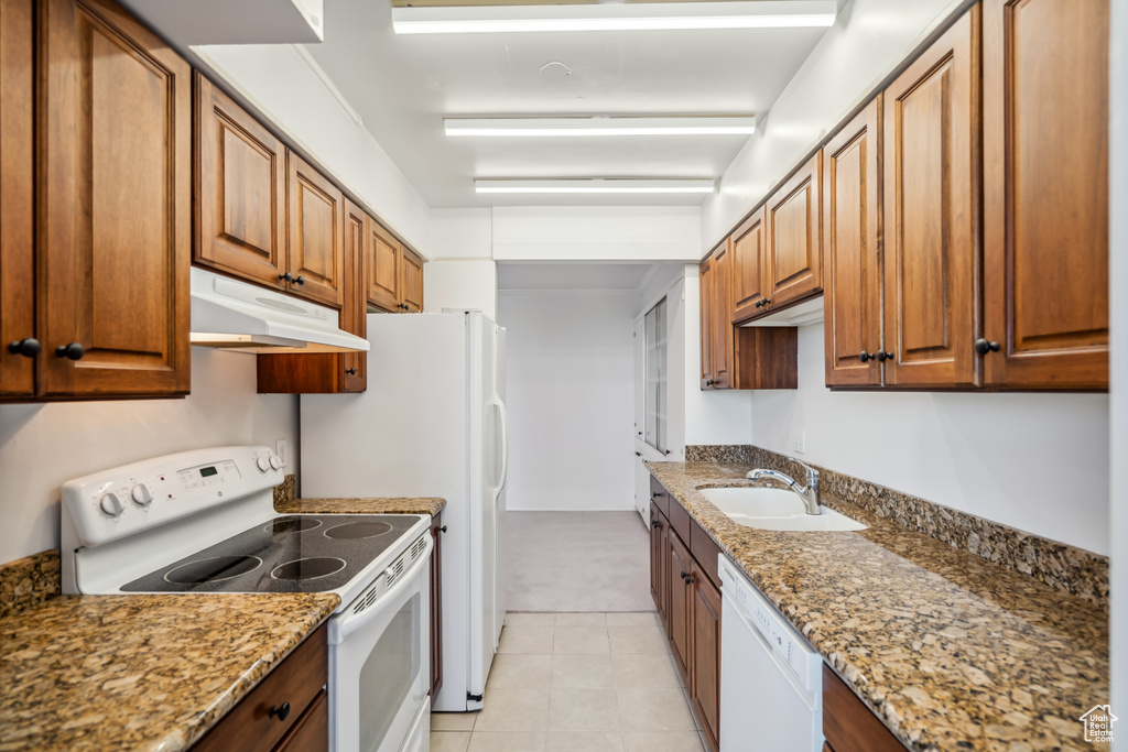 Kitchen featuring white appliances, under cabinet range hood, brown cabinetry, light stone counters, and light tile patterned floors