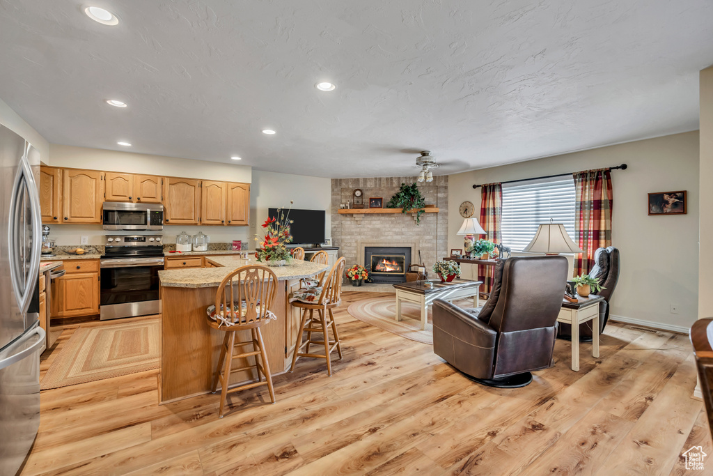 Kitchen with stainless steel appliances, light wood-style floors, recessed lighting, a kitchen breakfast bar, and a brick fireplace
