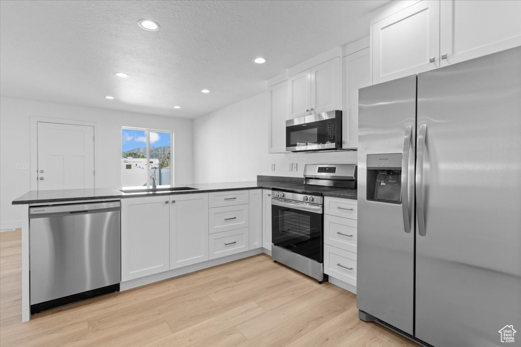 Kitchen with appliances with stainless steel finishes, a peninsula, light wood-type flooring, dark countertops, and recessed lighting