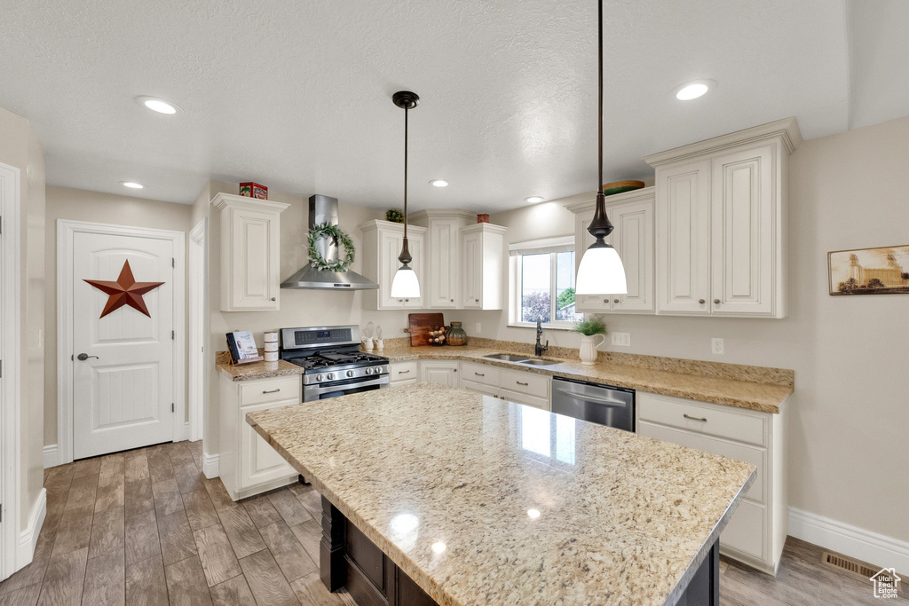 Kitchen featuring wall chimney exhaust hood, stainless steel appliances, light wood-type flooring, hanging light fixtures, and white cabinetry