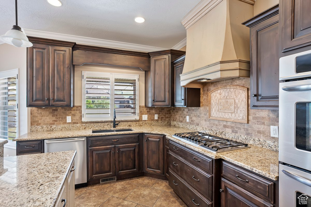 Kitchen featuring premium range hood, appliances with stainless steel finishes, dark brown cabinetry, backsplash, and ornamental molding
