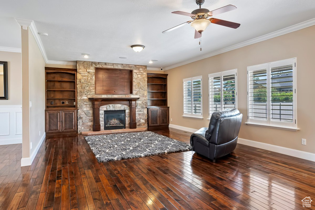 Living room featuring dark wood-type flooring, built in features, crown molding, a ceiling fan, and a fireplace