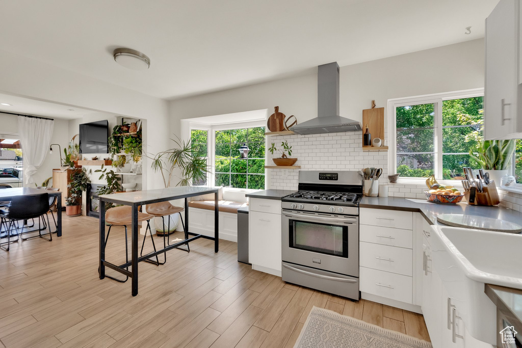 Kitchen featuring stainless steel range with gas stovetop, wall chimney range hood, light wood-type flooring, backsplash, and open shelves