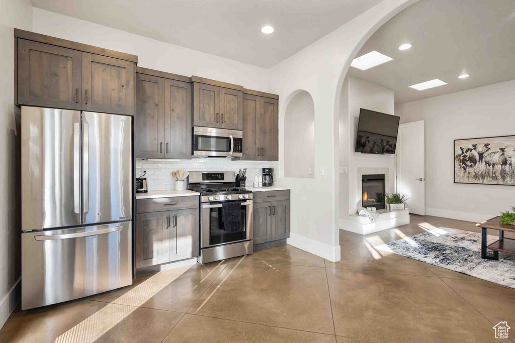 Kitchen featuring stainless steel appliances, arched walkways, tasteful backsplash, light countertops, and dark brown cabinets