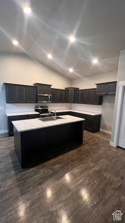 Kitchen featuring appliances with stainless steel finishes, dark wood finished floors, tasteful backsplash, a kitchen island with sink, and lofted ceiling