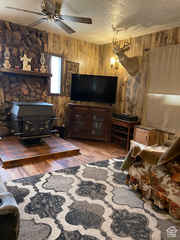 Living room with a wood stove, wooden walls, wood finished floors, a ceiling fan, and a textured ceiling
