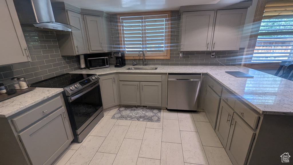 Kitchen featuring stainless steel appliances, gray cabinets, wall chimney exhaust hood, tasteful backsplash, and light stone countertops