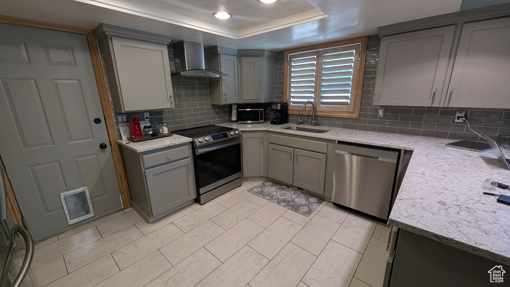 Kitchen featuring stainless steel appliances, wall chimney range hood, gray cabinetry, backsplash, and a raised ceiling