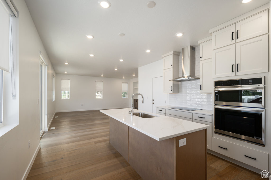Kitchen with double oven, wall chimney exhaust hood, decorative backsplash, a center island with sink, and white cabinets
