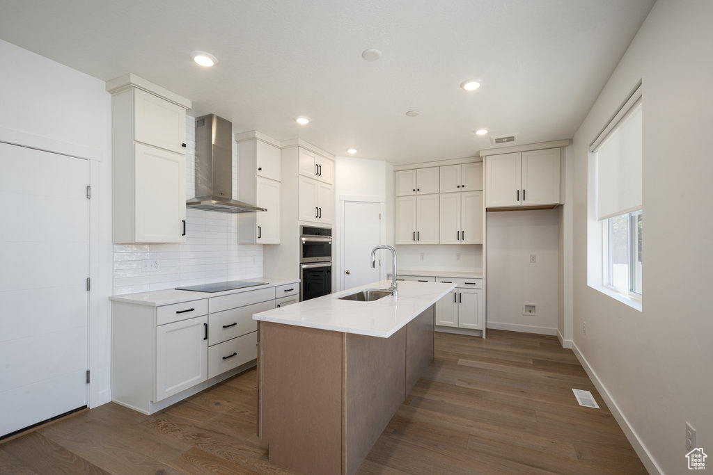Kitchen featuring wall chimney range hood, black electric cooktop, backsplash, double oven, and white cabinetry