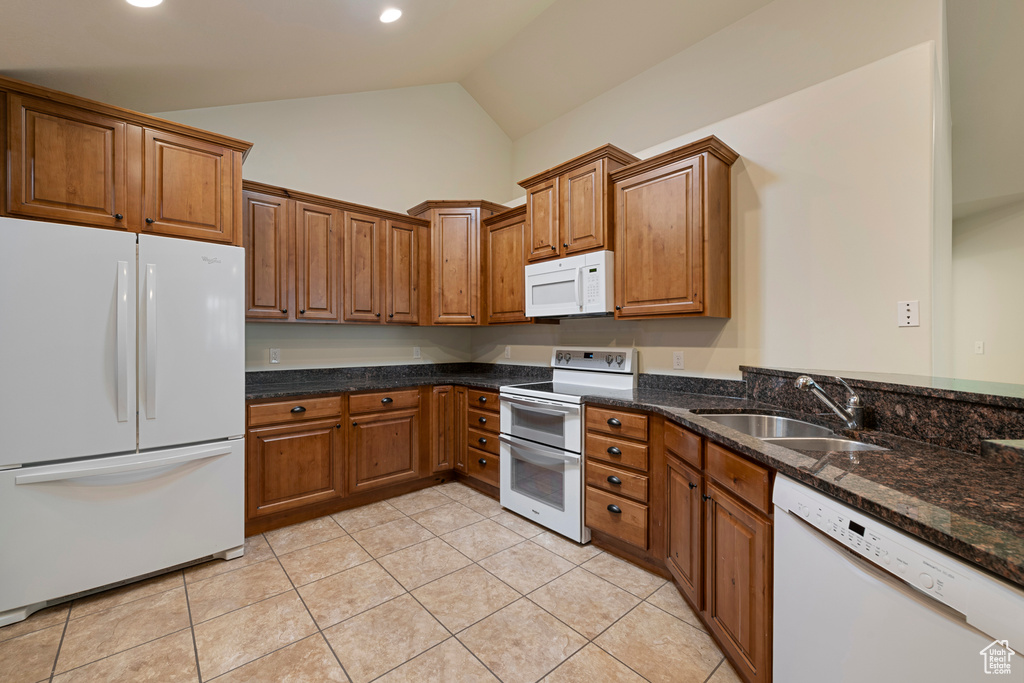 Kitchen featuring white appliances, brown cabinetry, light tile patterned flooring, dark stone counters, and high vaulted ceiling