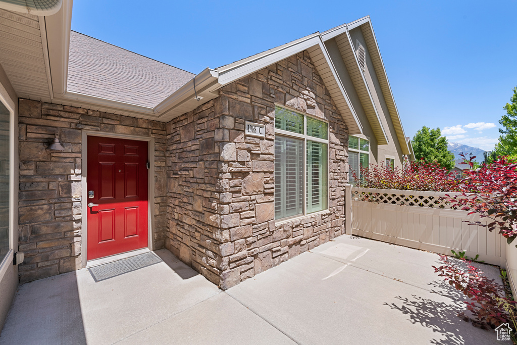 View of exterior entry featuring stone siding, a shingled roof, and a patio