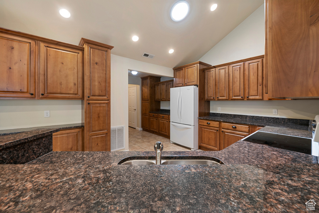 Kitchen featuring freestanding refrigerator, lofted ceiling, brown cabinetry, dark stone countertops, and electric range oven