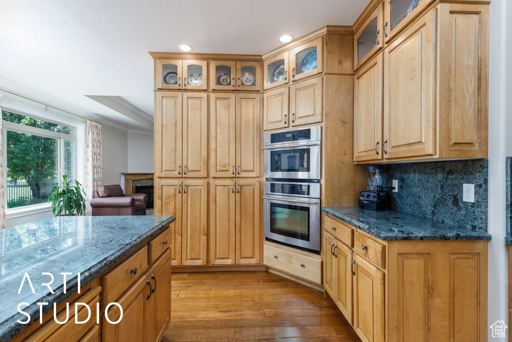 Kitchen featuring double oven, wood-type flooring, tasteful backsplash, dark stone countertops, and glass insert cabinets