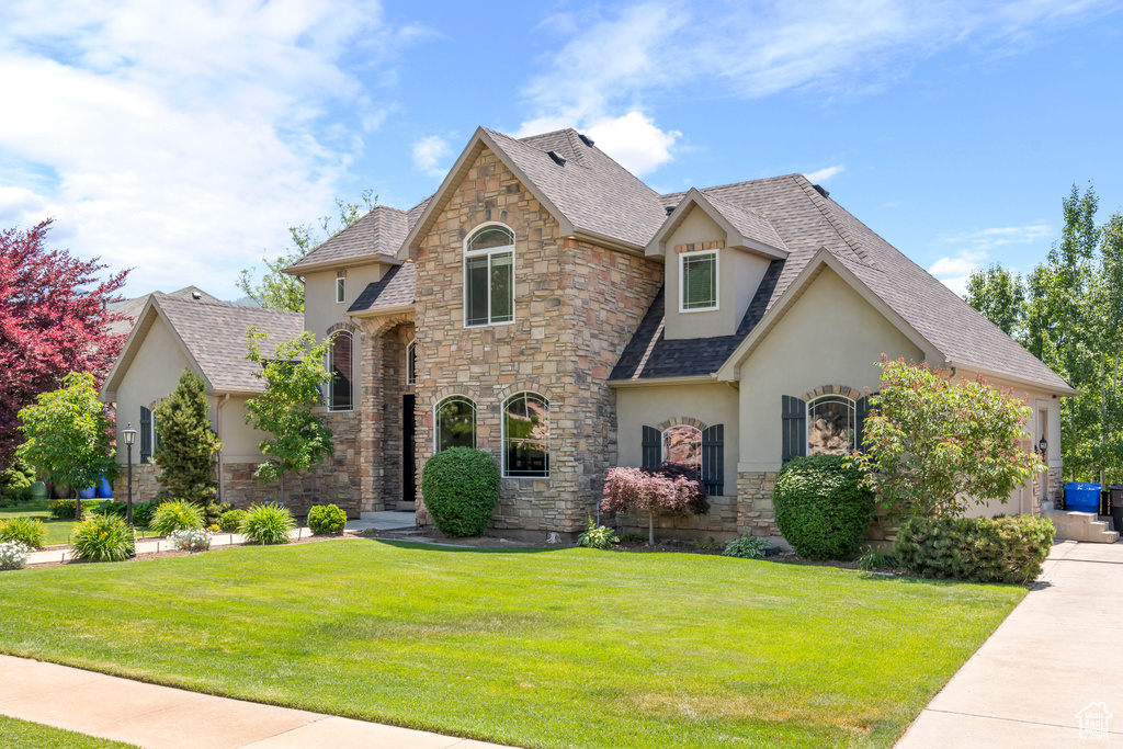French provincial home featuring stucco siding, stone siding, a front yard, and roof with shingles